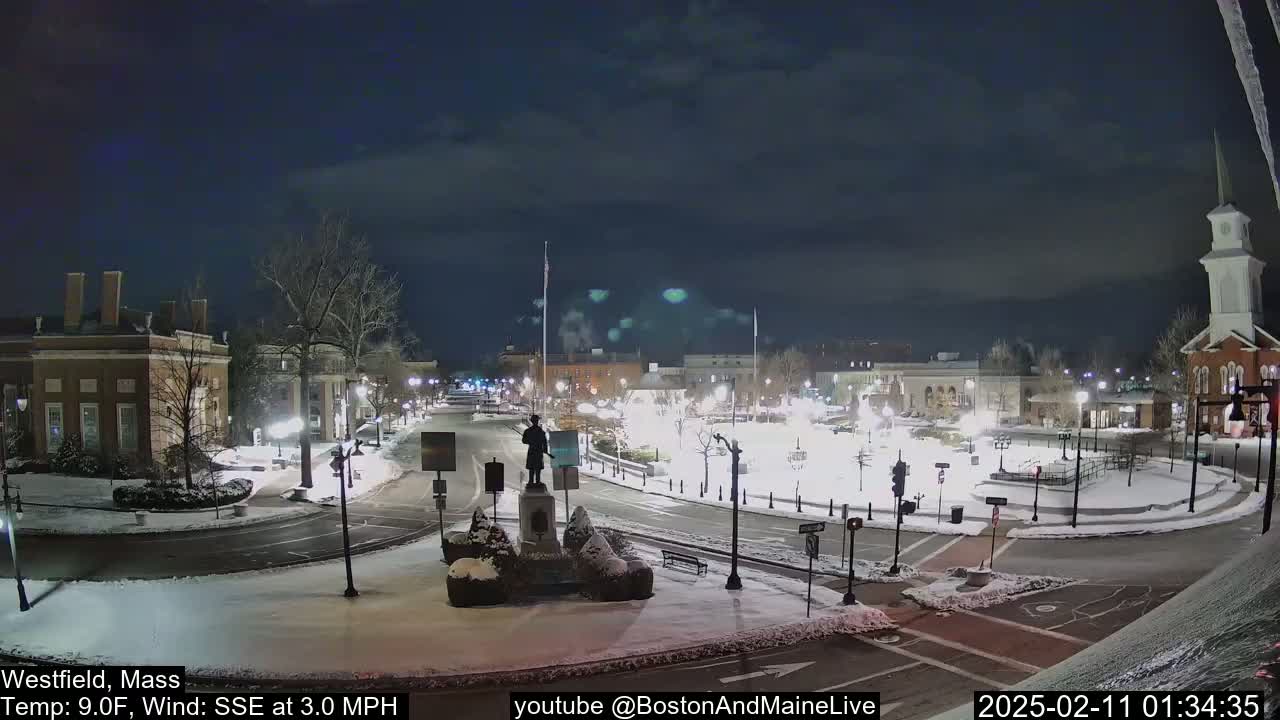 A snow-covered town square at night, illuminated by streetlights, features a statue in the center and buildings surrounding it under a partly cloudy sky.