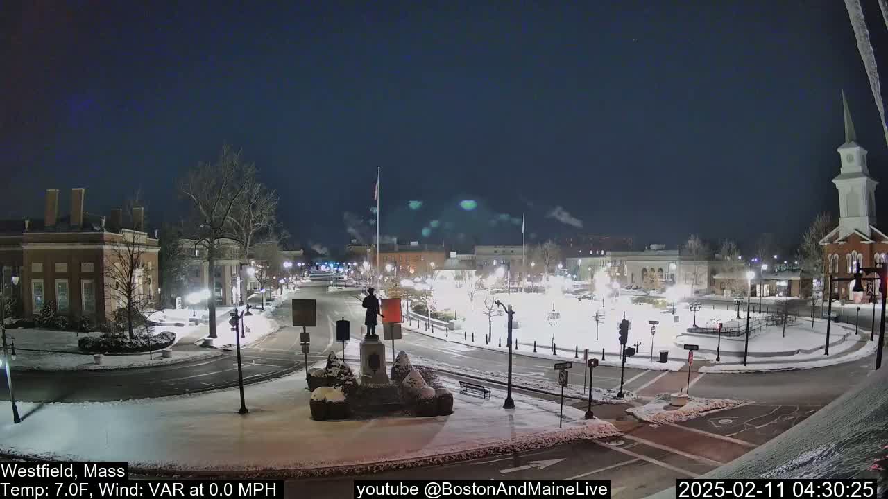 A snow-covered town square at night, illuminated by streetlights, features a statue in the center and buildings surrounding it under a clear, dark sky.