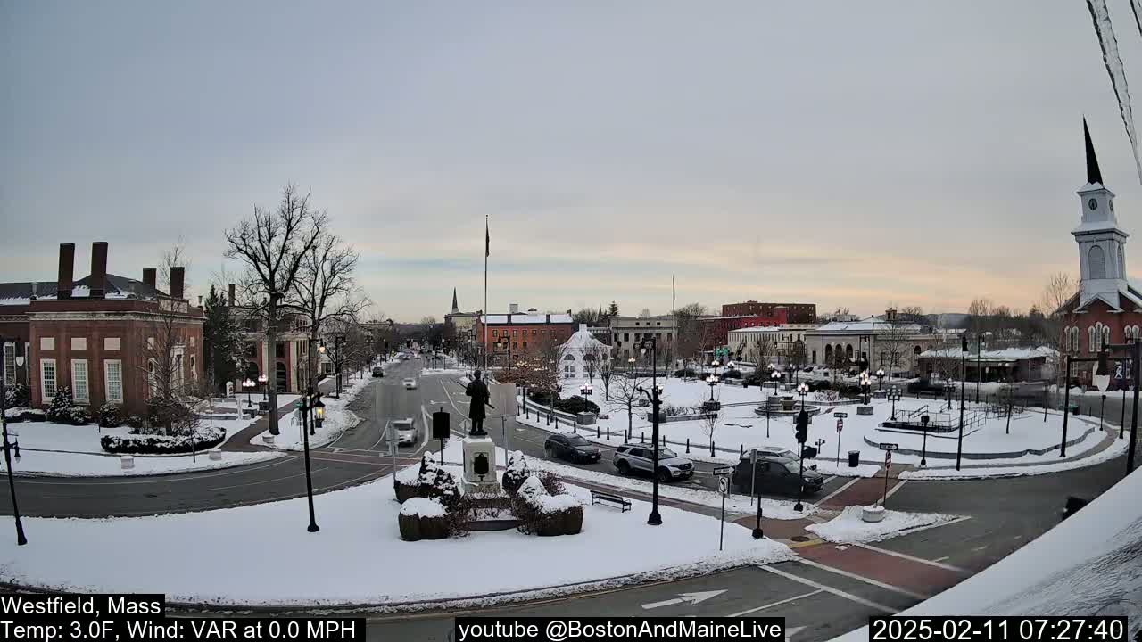 A snow-covered town square with brick buildings and a statue in the center, under a cloudy but clear sky.
