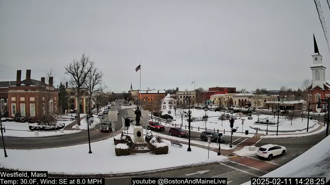 A snow-covered town square with several brick buildings, a statue, and cars driving around a traffic circle under an overcast sky.