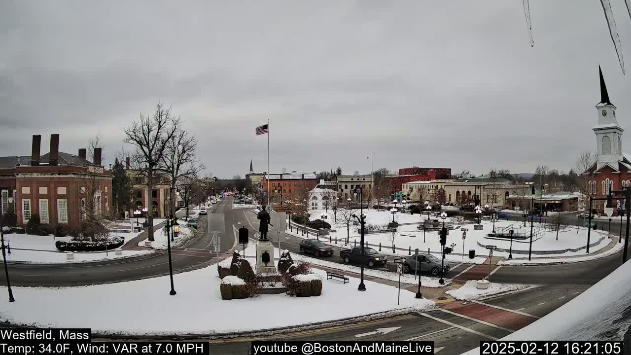 A snow-covered town square with brick buildings, a statue, and several cars, under an overcast sky.