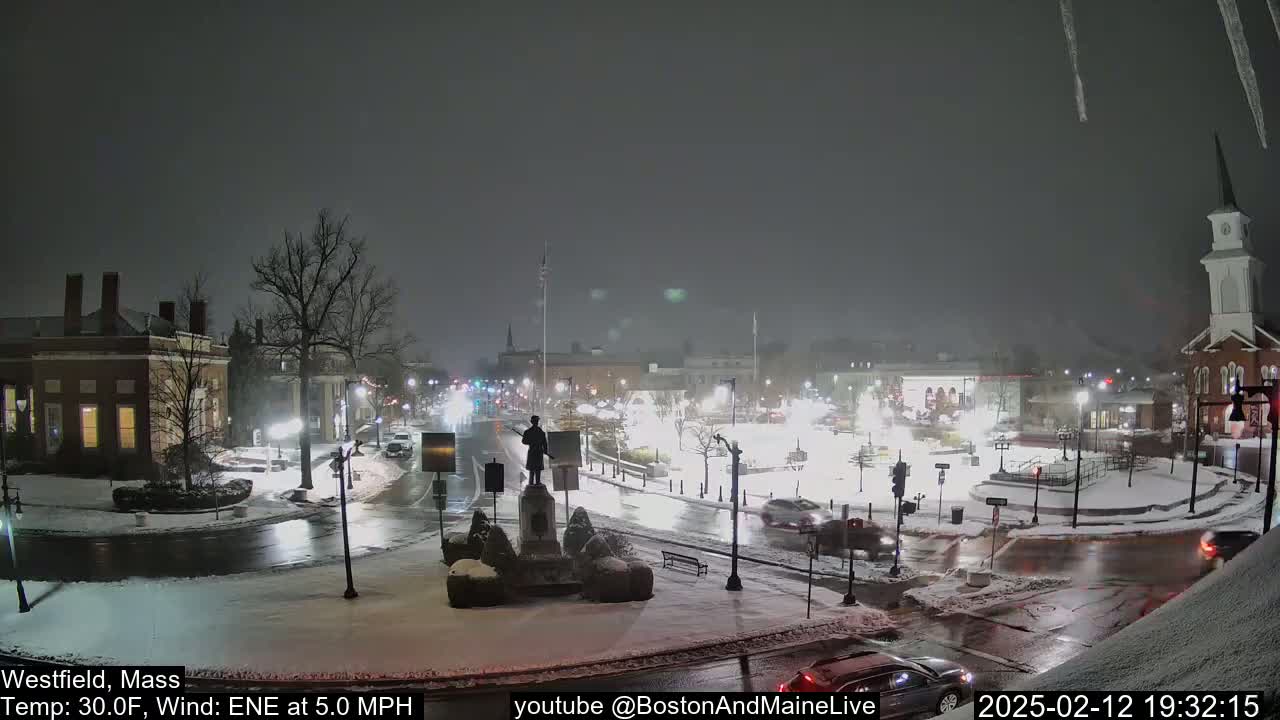 A snow-covered town square at night, with cars driving on wet roads and a statue in the center, under a lightly falling snow.