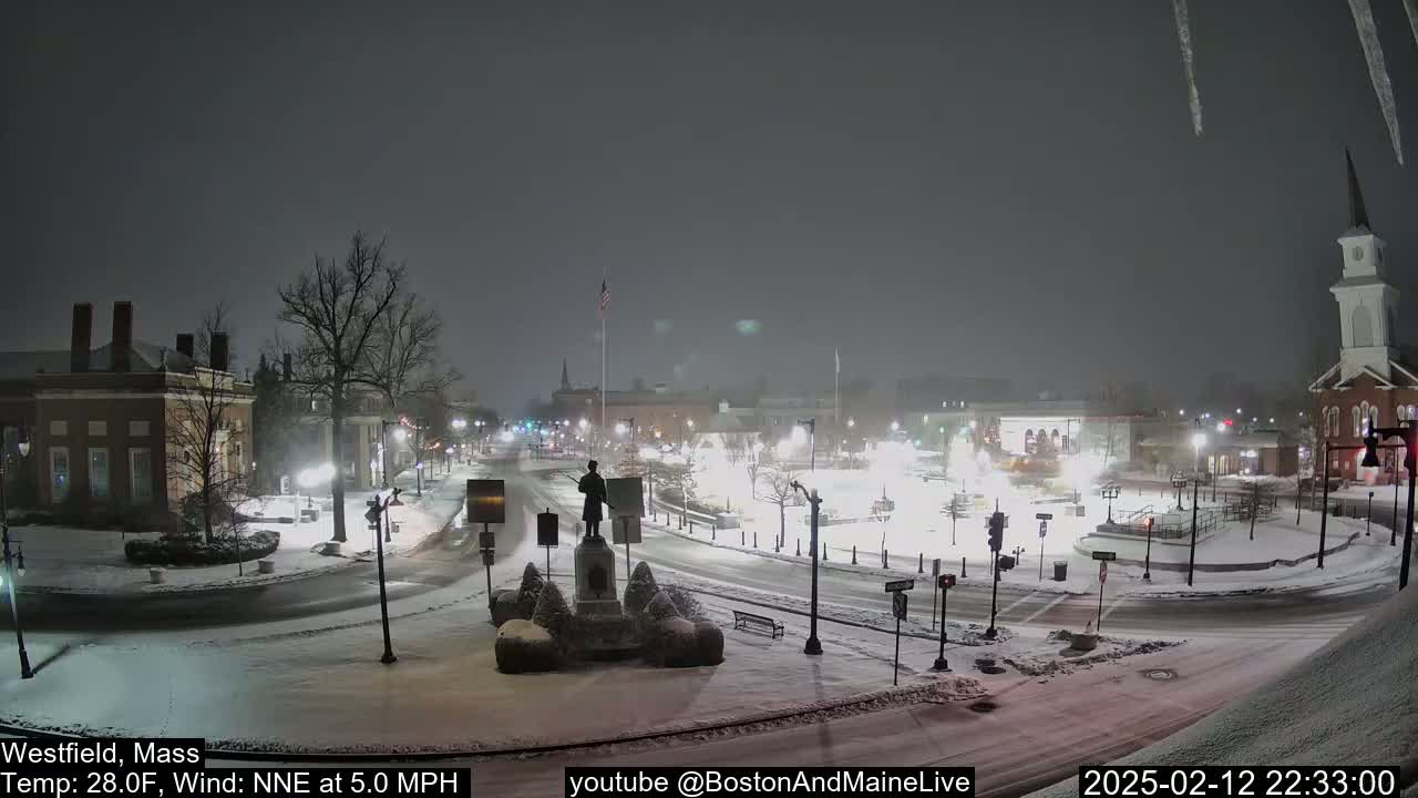 A snow-covered town square at night, featuring a statue in the center, is lightly dusted with snow under a dark, overcast sky.
