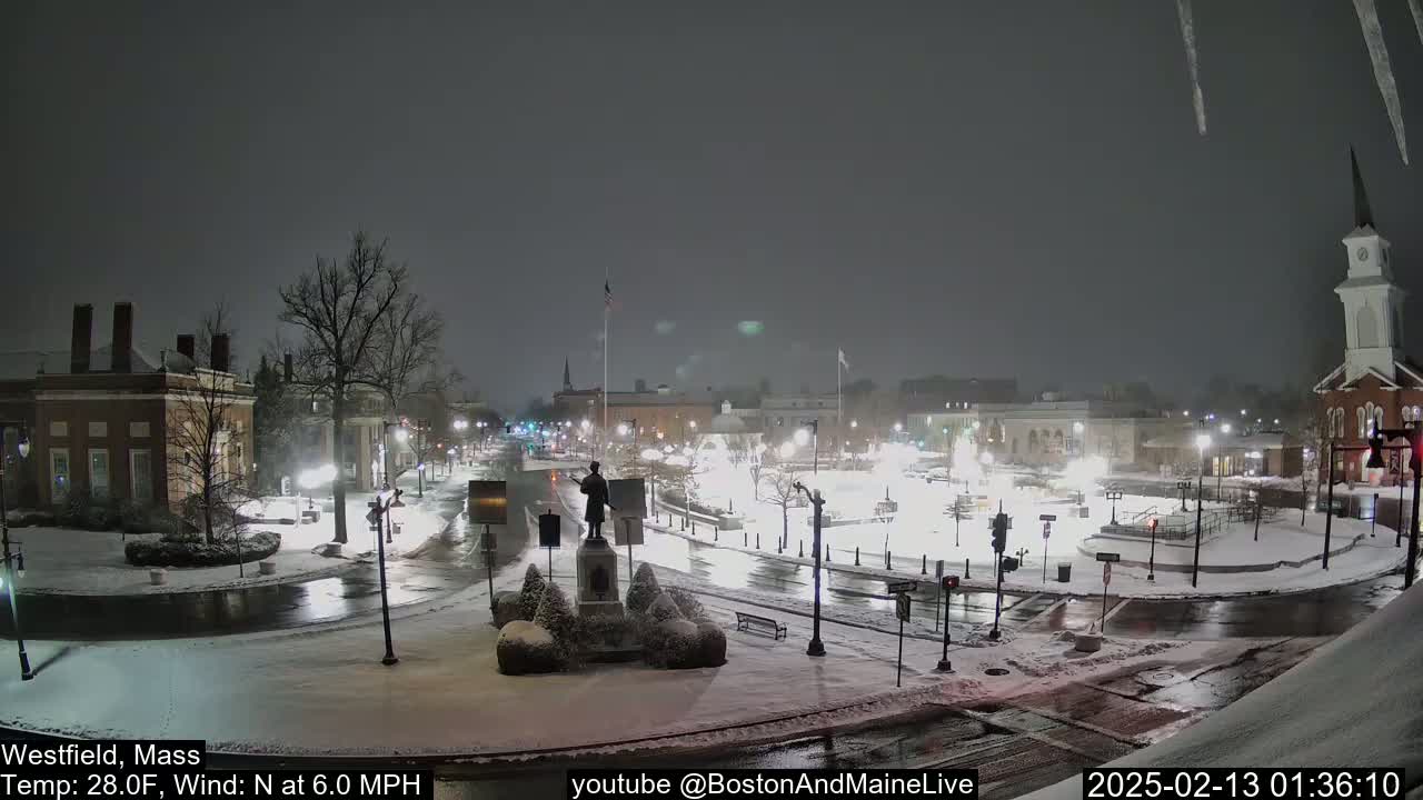 A snow-covered town square at night, lightly snowing with a north wind, features a statue in the center and buildings surrounding it.