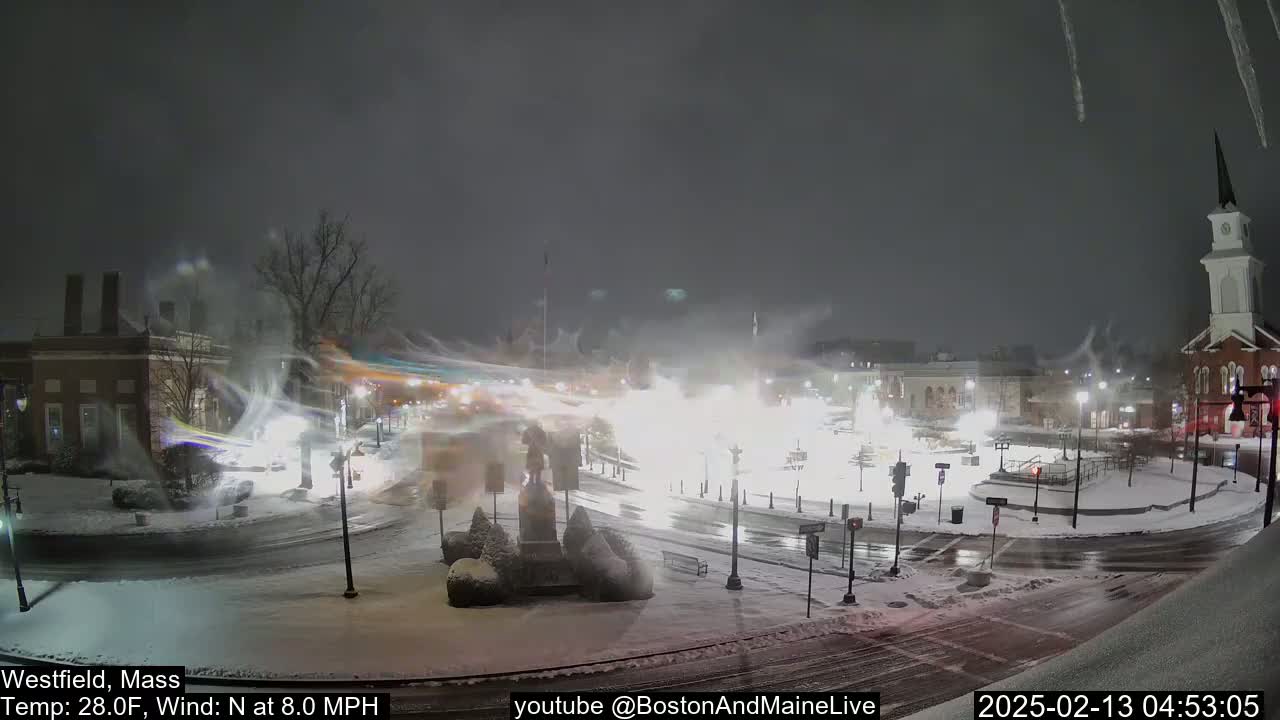A snow-covered town square at night, with a church steeple visible in the background, under windy, snowy conditions.