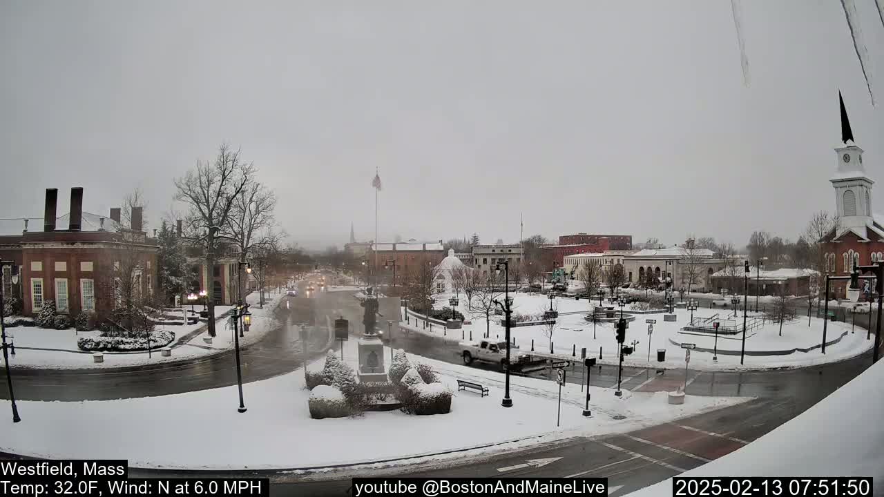 A snow-covered town square, with cars driving on wet roads and buildings surrounding it under an overcast sky.