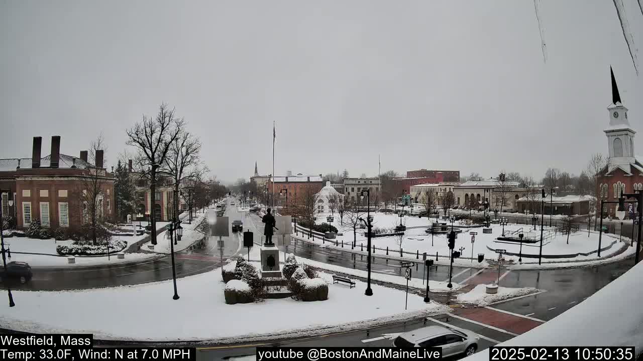 A snow-covered town square with brick buildings, a statue, and a church steeple under a gray sky.