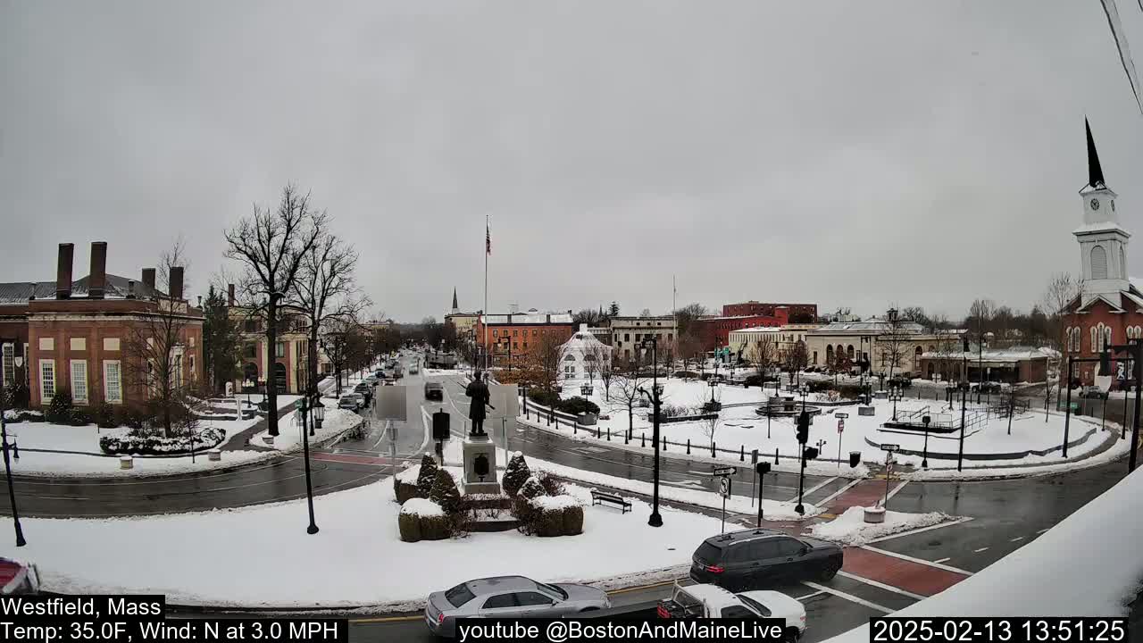 A snow-covered town square with brick buildings, a statue, and cars, under an overcast sky.