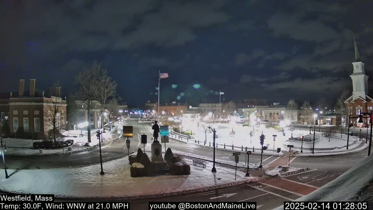 A snow-covered town square at night, illuminated by streetlights, features a statue in the center and several buildings surrounding it under a partly cloudy sky.
