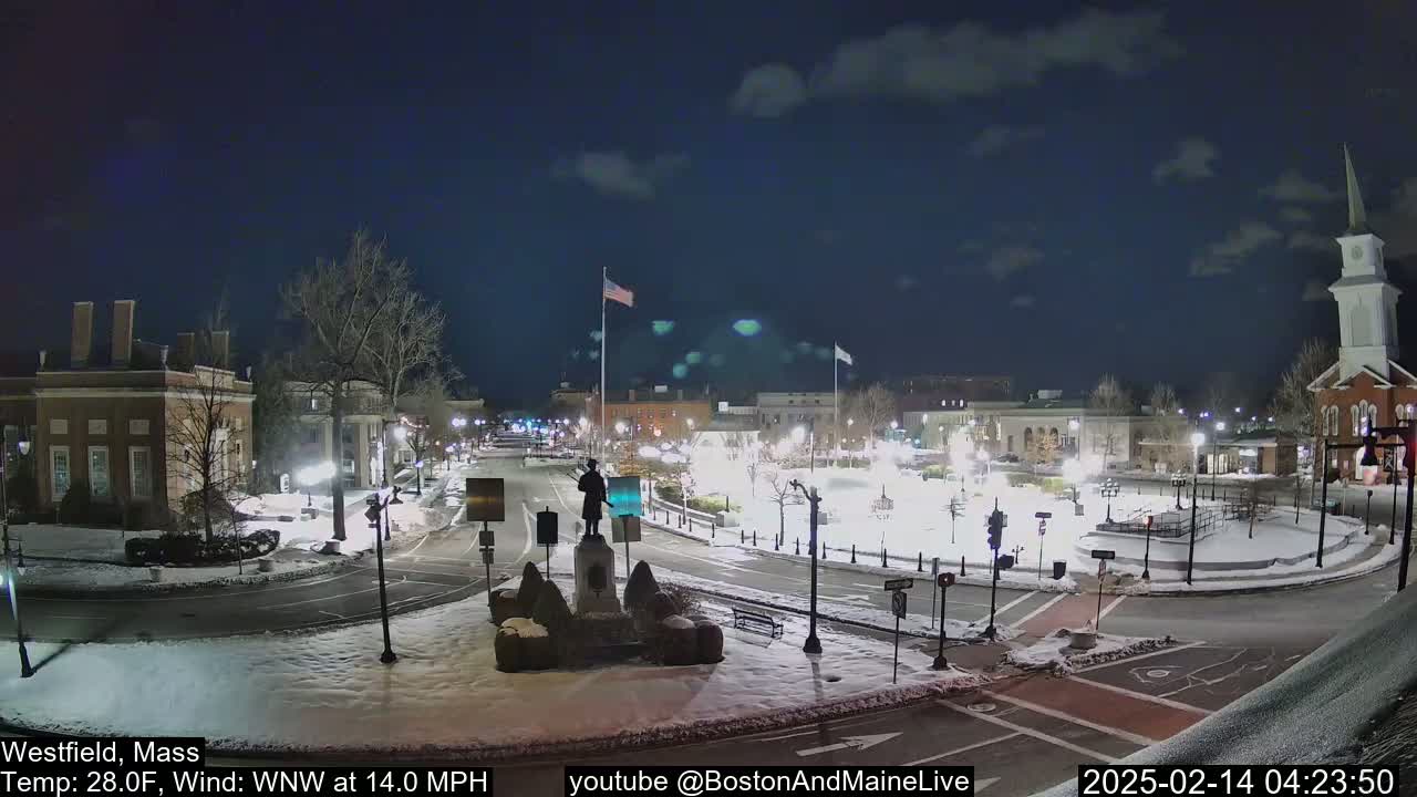 A snow-covered town square at night, illuminated by streetlights, with a statue in the center and buildings surrounding it under a partly cloudy sky.