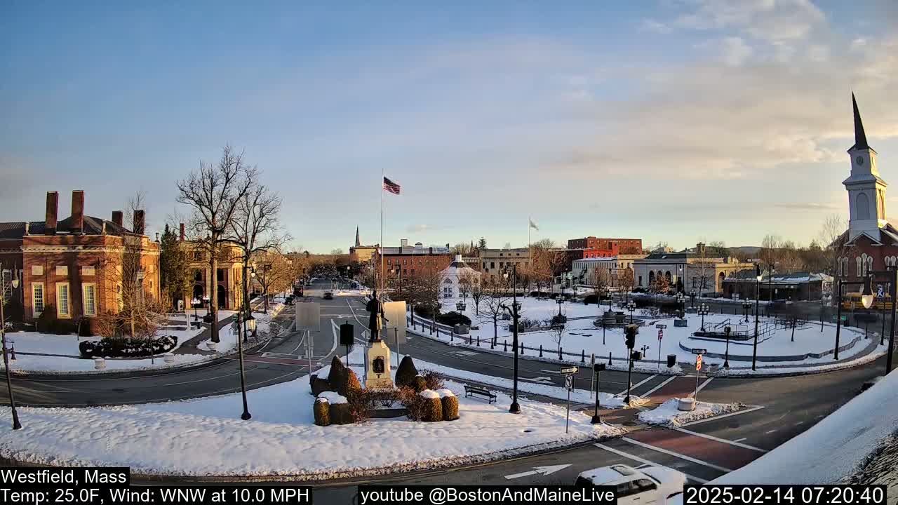 A snow-covered town square with brick buildings, a monument, and a church steeple under a partly cloudy sky.