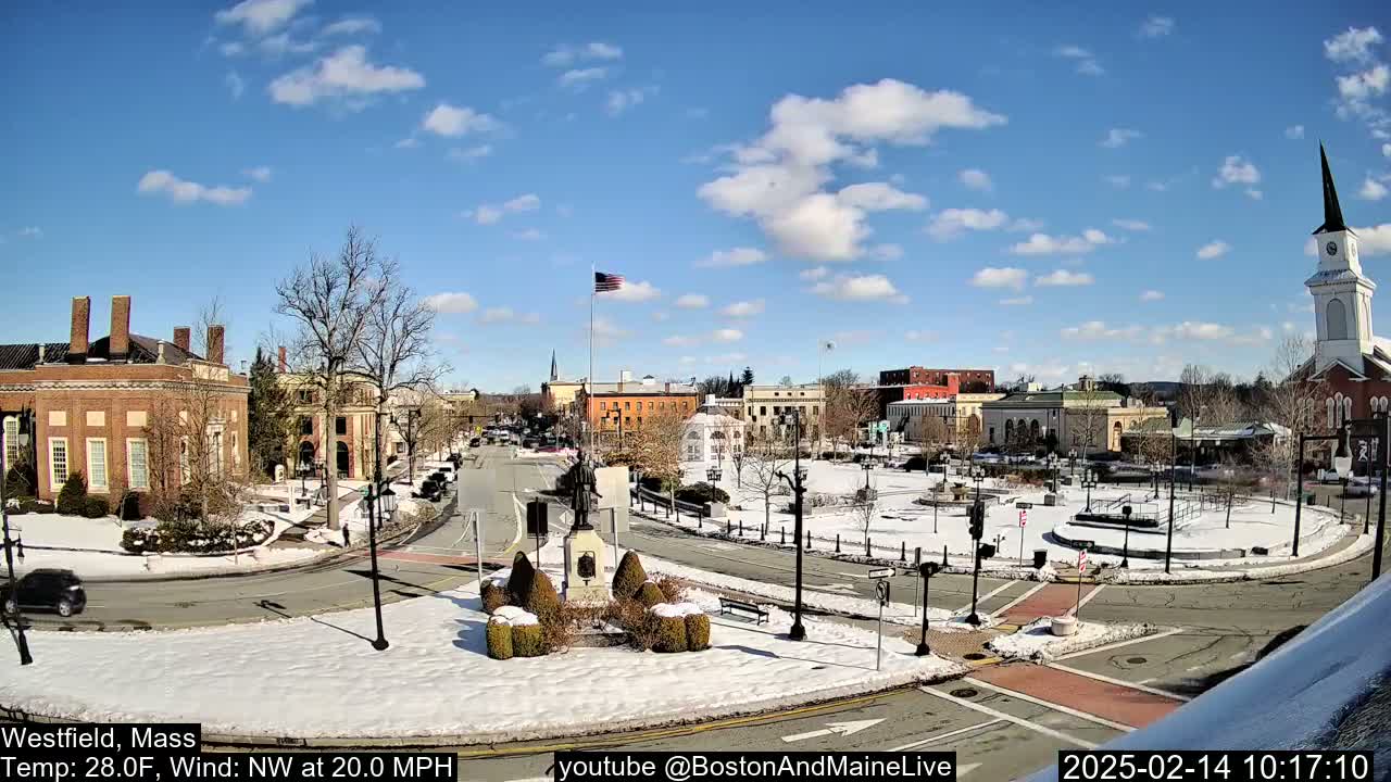 A snow-covered town square with brick buildings, a statue, and a church steeple under a partly cloudy blue sky.