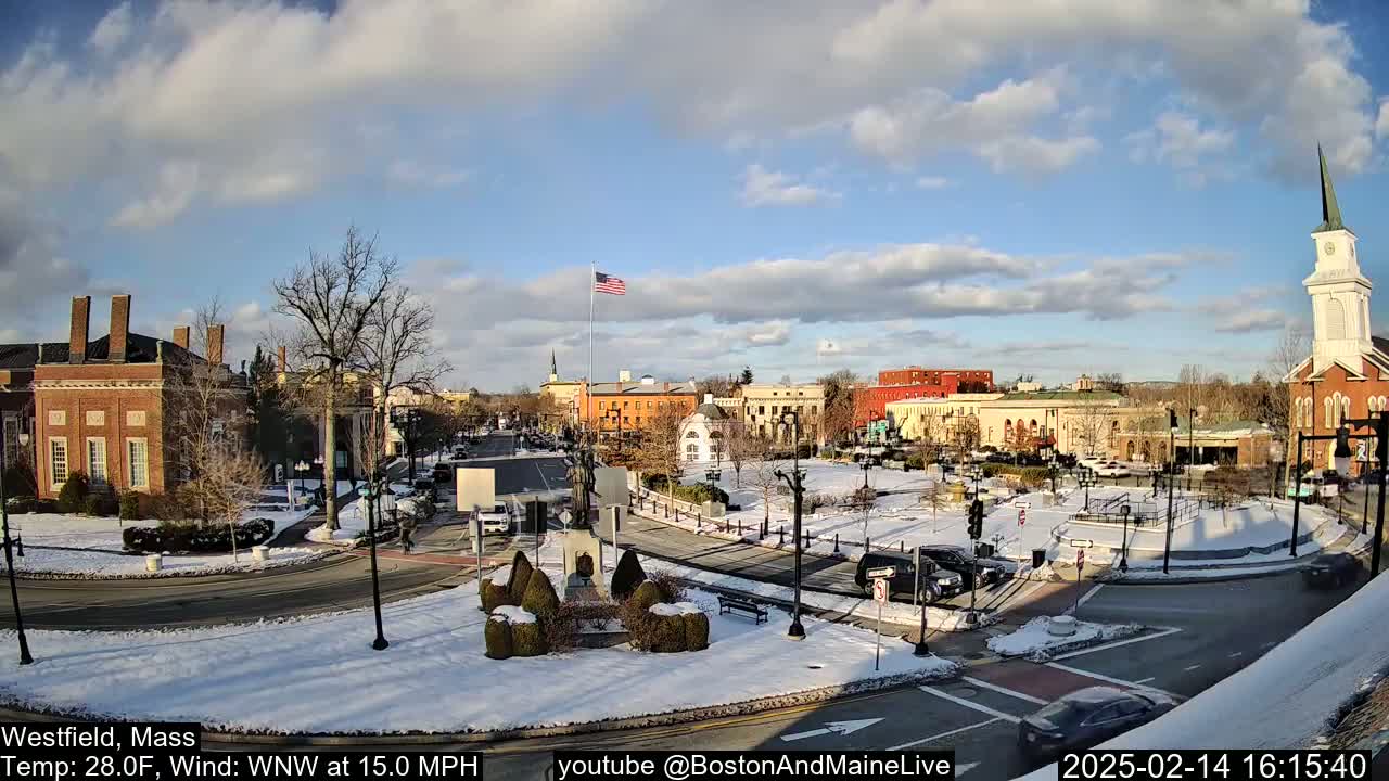 A snow-covered town square with brick buildings, a church steeple, and a few cars, under a partly cloudy sky.