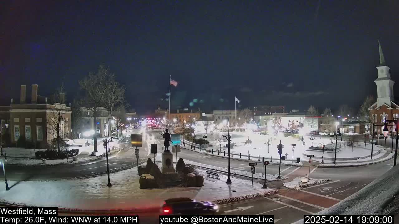 A snow-covered town square at night, illuminated by streetlights, with a statue in the center and buildings surrounding it, under a clear dark sky with a 14 mph wind.