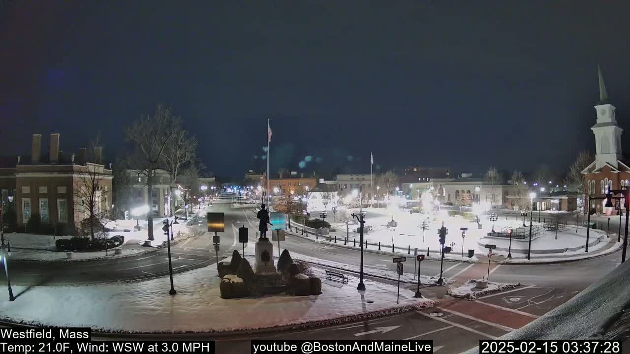 A snow-covered town square at night, illuminated by streetlights, features a statue in the center and several buildings surrounding it.