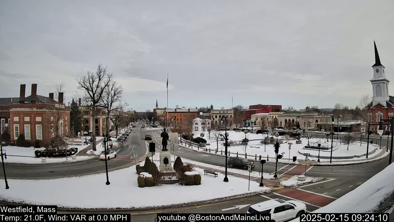 A snow-covered town square with a statue in the center, surrounded by brick buildings and a church steeple, under a cloudy sky.