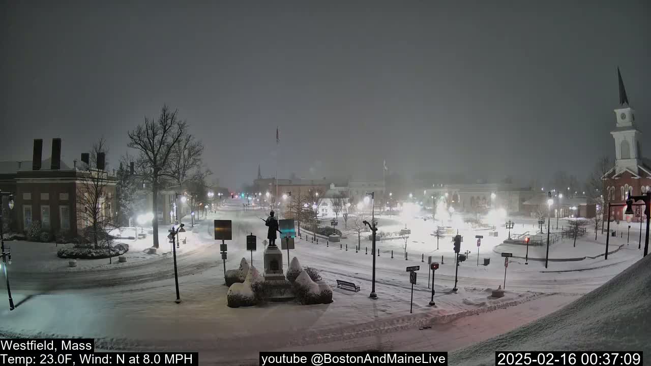 A snow-covered town square at night, featuring a statue, streetlights, and buildings, under falling snow with a north wind.