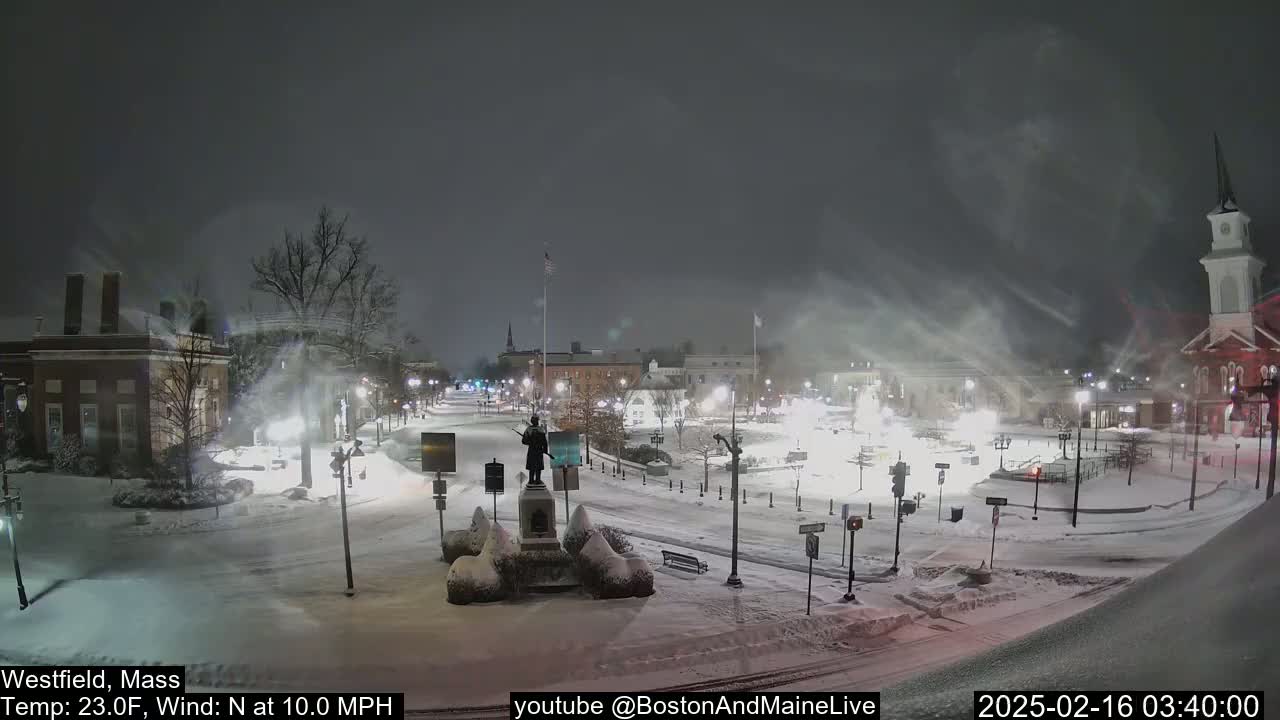 A snow-covered town square at night with a statue in the center, under blowing snow and a 23°F temperature.