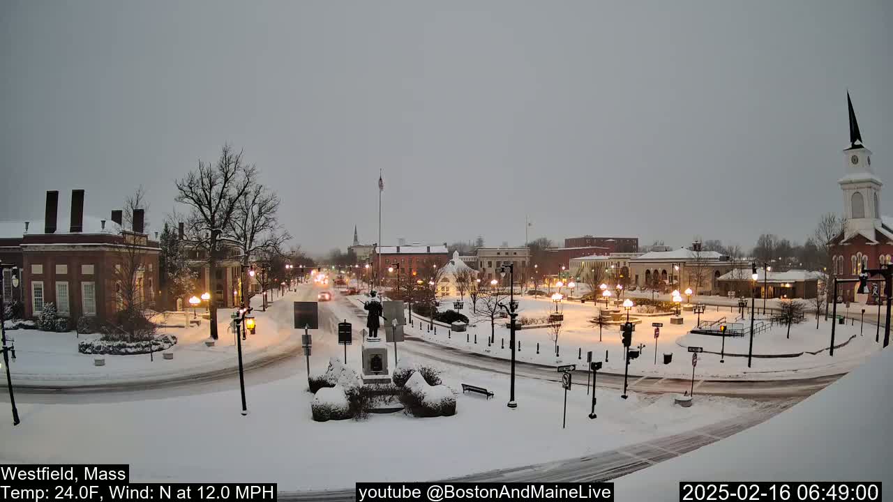 A snow-covered town square, with brick buildings and a church steeple, is lightly trafficked under a gray, overcast sky.
