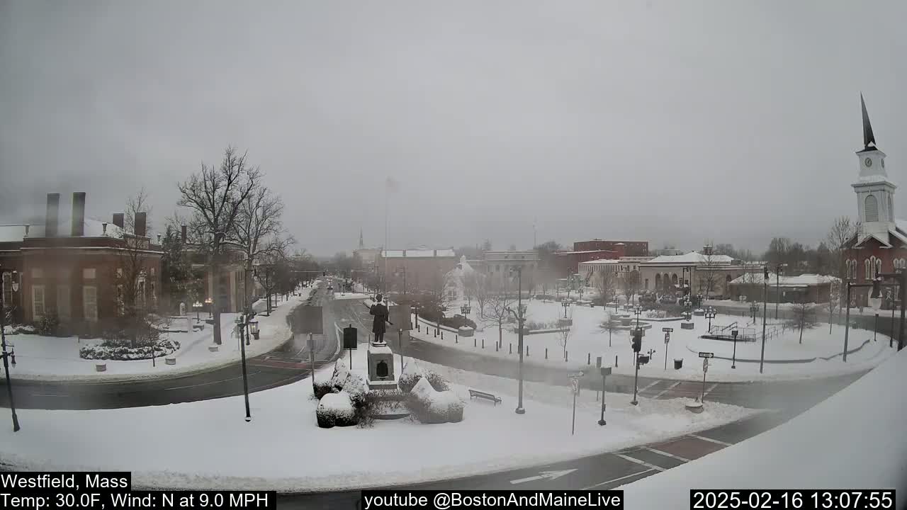 A snow-covered town square with brick buildings, a statue, and a church steeple is visible under a gray, overcast sky.