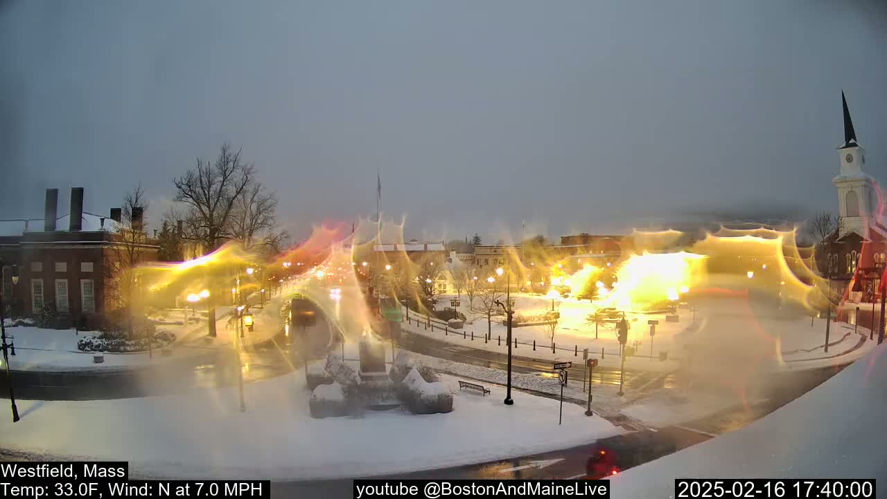 A snow-covered town square at dusk, illuminated by streetlights, with a church steeple in the background and a 7 mph north wind.