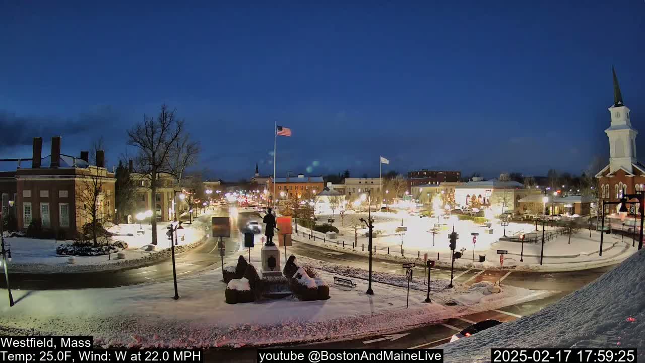 A snow-covered town square at night, illuminated by streetlights, with a statue in the center and a church steeple visible in the background, under a clear, dark blue sky.