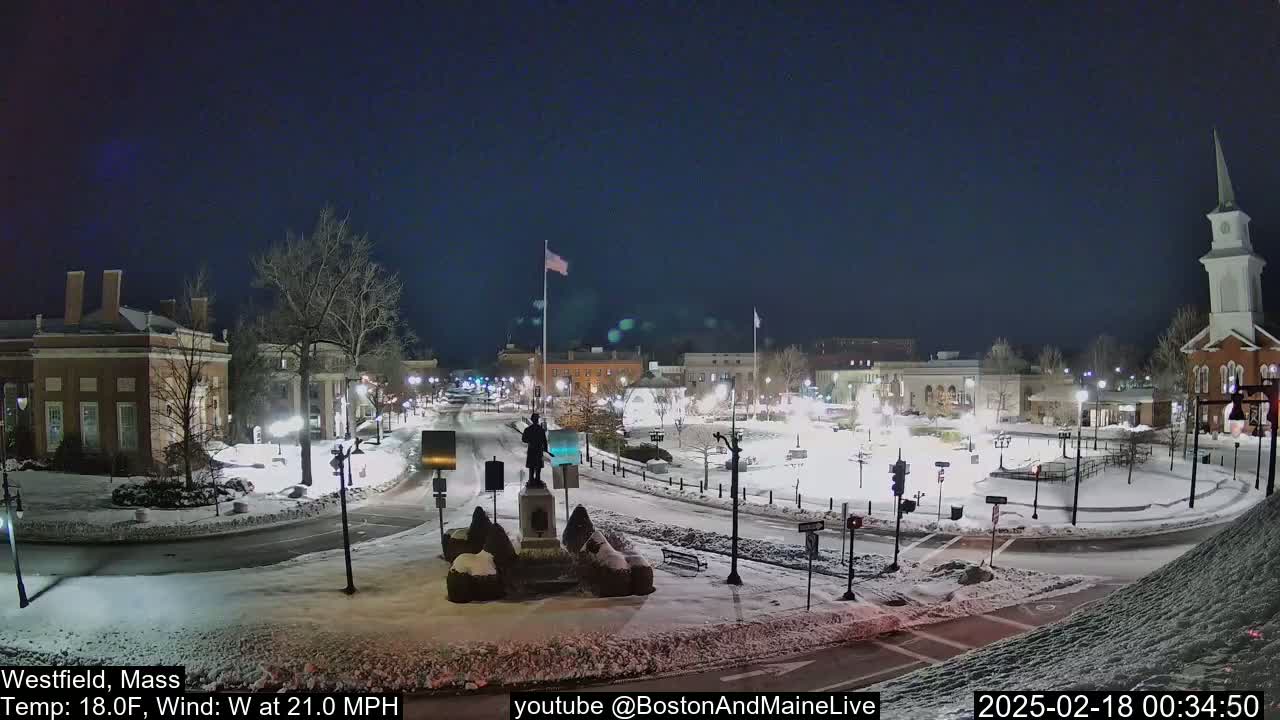 A snow-covered town square at night, with a statue in the center, under a dark sky, and a wind of 21 mph.