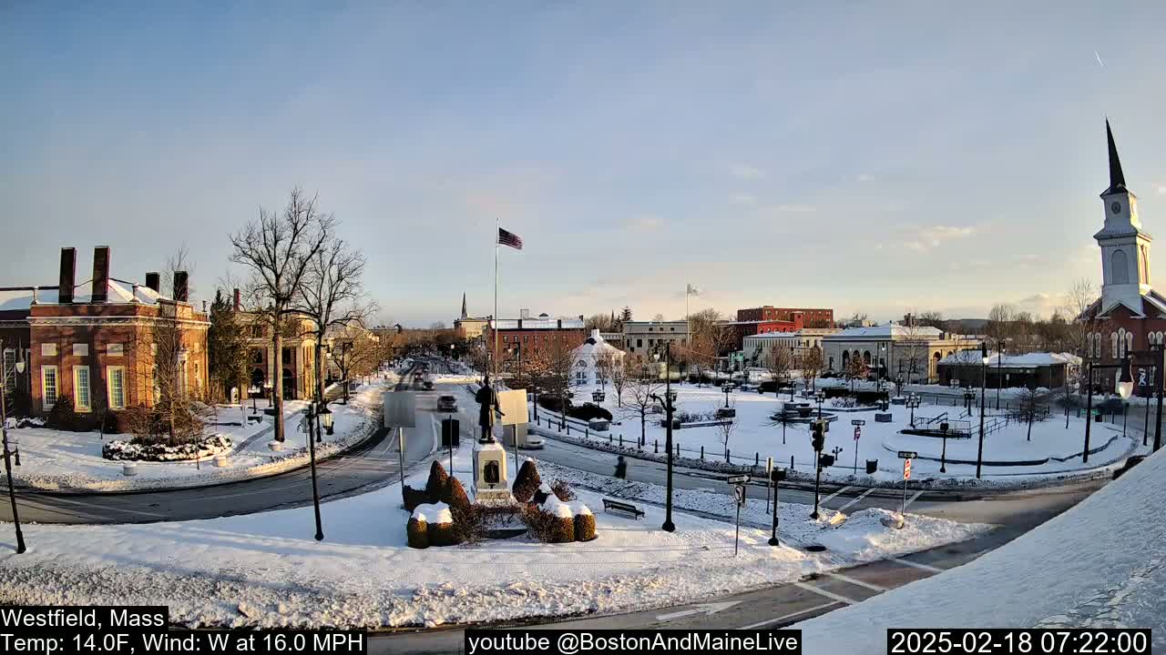 A snow-covered town square with brick buildings, a monument, and a church steeple is visible under a clear, bright sky.
