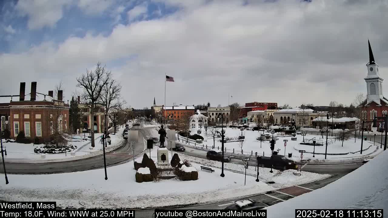 A snow-covered town square with brick buildings, a monument, and a church steeple under a partly cloudy sky, with a strong wind.