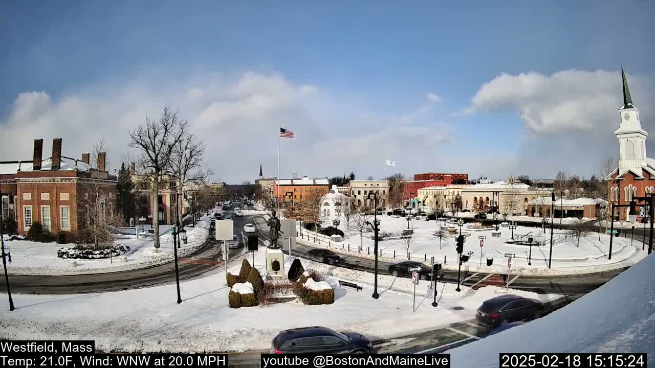 A snow-covered town square with brick buildings, a monument, and cars driving on roads under a partly cloudy sky.