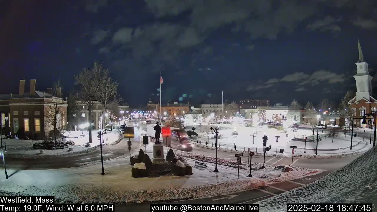 A snow-covered town square at night, illuminated by streetlights, features a statue in the center and several buildings surrounding it, with a church steeple visible in the background under a partly cloudy sky.