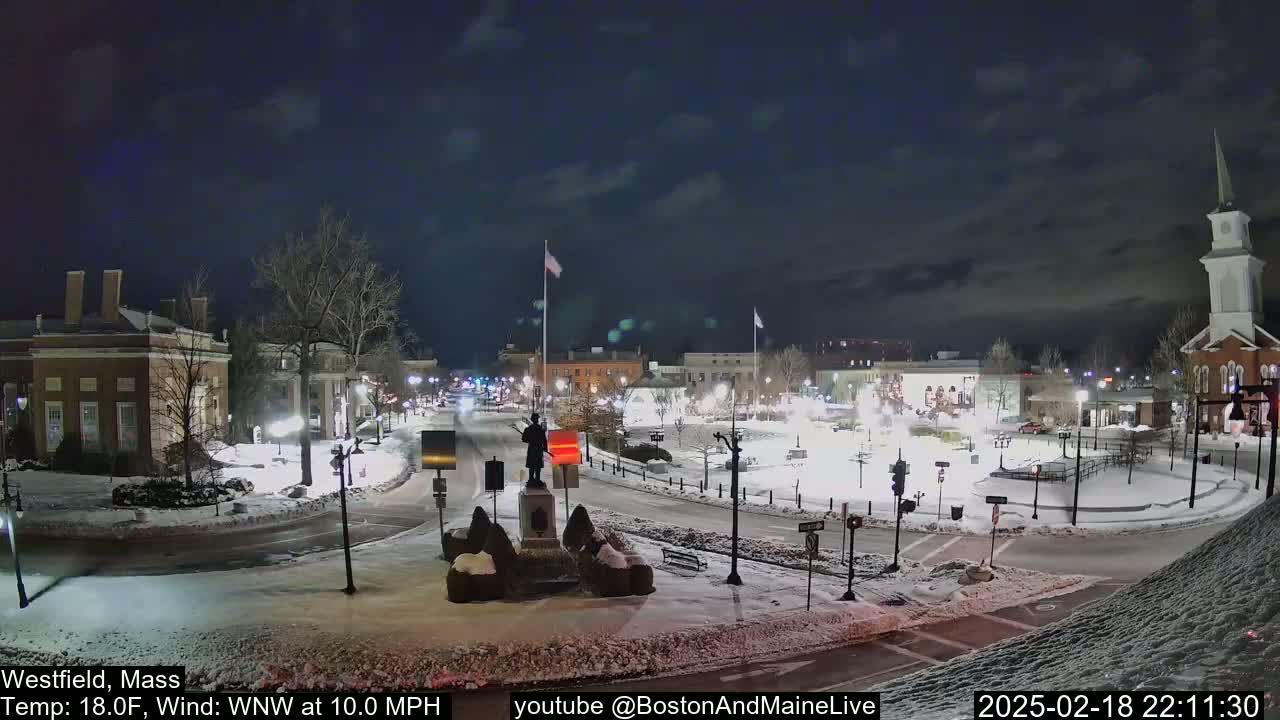A snow-covered town square at night, illuminated by streetlights, features a statue in the center and buildings surrounding the perimeter under a partly cloudy sky.