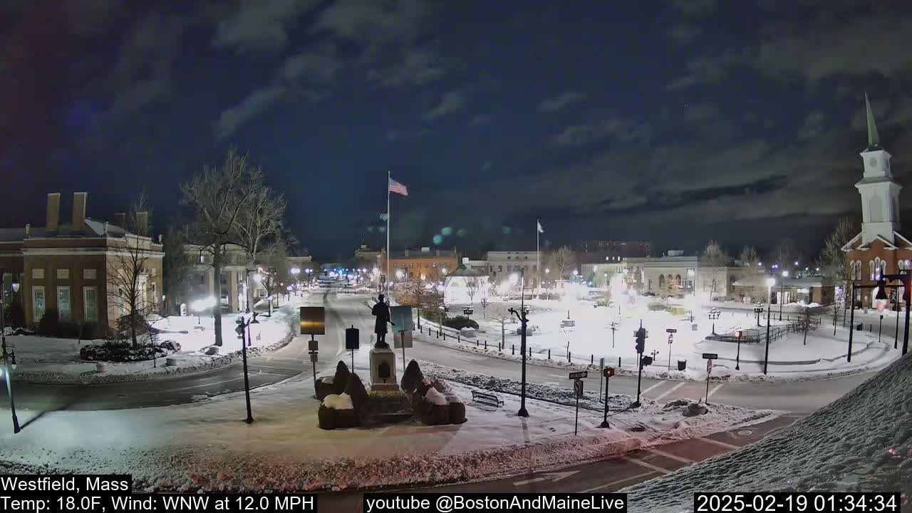 A snow-covered town square at night, illuminated by streetlights, features a statue in the center, with buildings surrounding it under a partly cloudy sky.