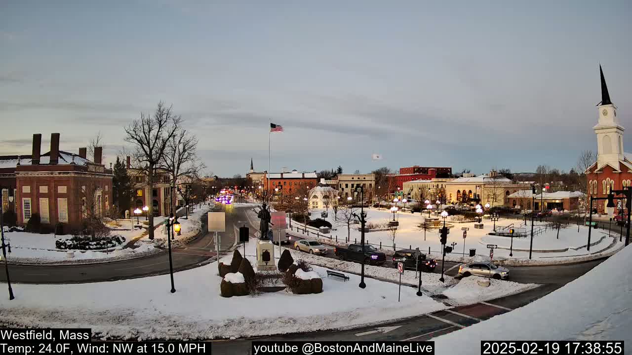 A snow-covered town square with brick buildings, a statue, and a church steeple under a twilight sky, with a northwest wind at 15 mph.