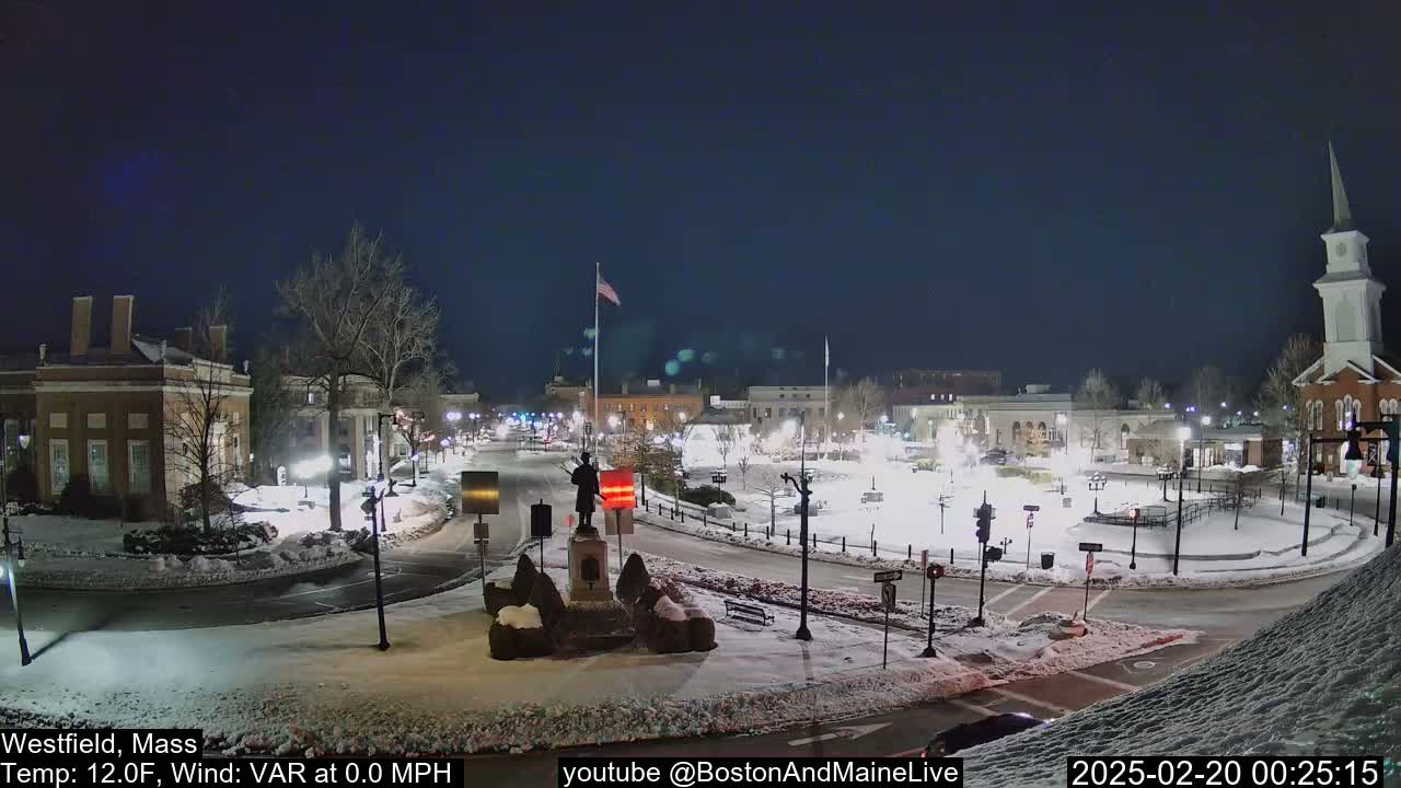 A snow-covered town square at night, illuminated by streetlights, features a statue in the center, surrounded by buildings and a church steeple.