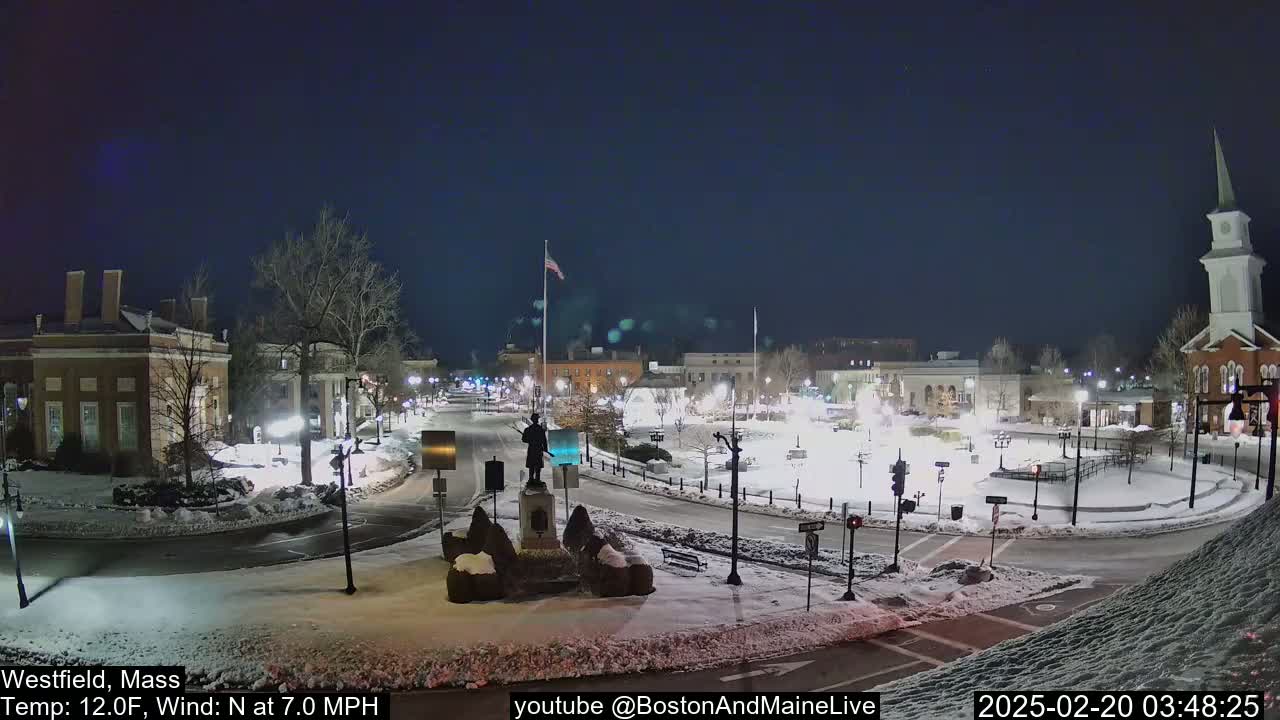 A snow-covered town square at night, illuminated by streetlights, features a statue in the center and buildings surrounding it, with a church steeple visible in the background under a clear, dark sky.