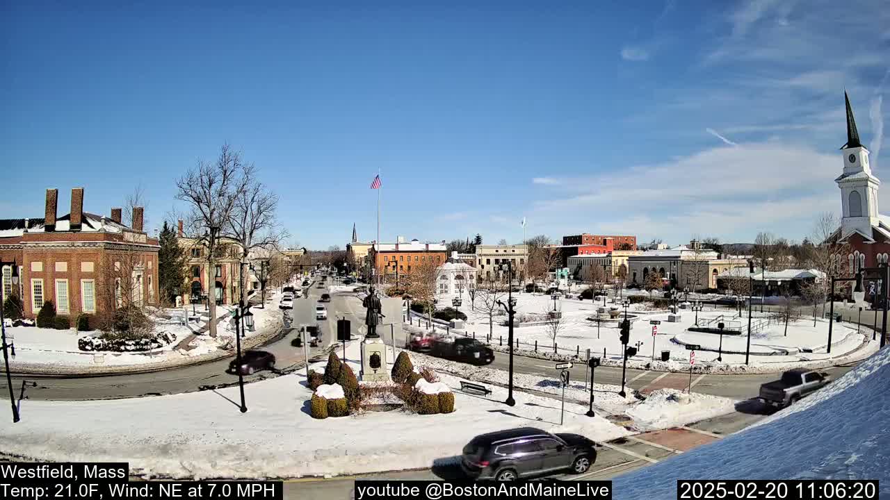 A snow-covered town square with brick buildings, a statue, and cars driving around a traffic circle under a clear blue sky.