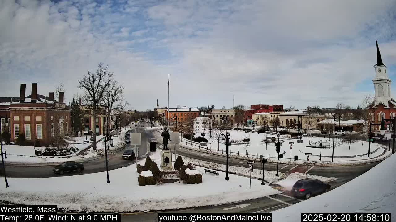 A snow-covered town square with brick buildings, a statue, and a church steeple under a partly cloudy sky.