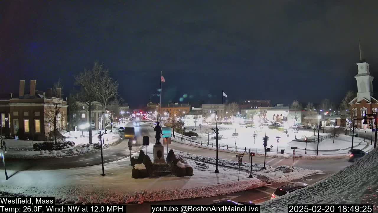 A snow-covered town square at night, illuminated by streetlights, features a statue in the center, with a few cars visible on the roads.