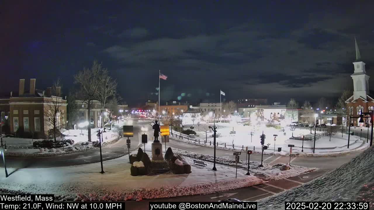 A snow-covered town square at night, featuring a statue, buildings, and streetlights under a partly cloudy sky with a northwest wind.