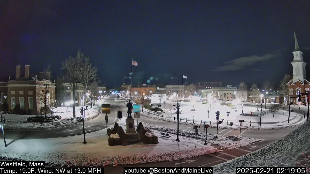 A snow-covered town square at night, illuminated by streetlights, features a statue in the center and buildings surrounding it under a mostly clear sky.