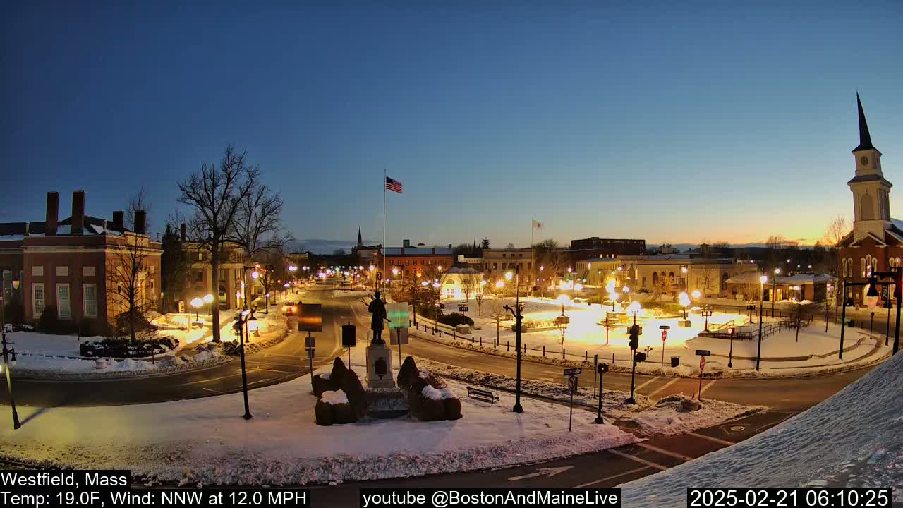 A snow-covered town square at twilight, illuminated by streetlights, features a statue in the center and buildings surrounding it, with a church steeple in the background under a clear, dark blue sky.