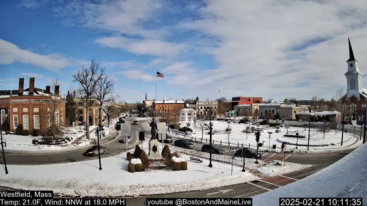 A snow-covered town square with brick buildings, a monument, and a church steeple under a partly sunny sky.