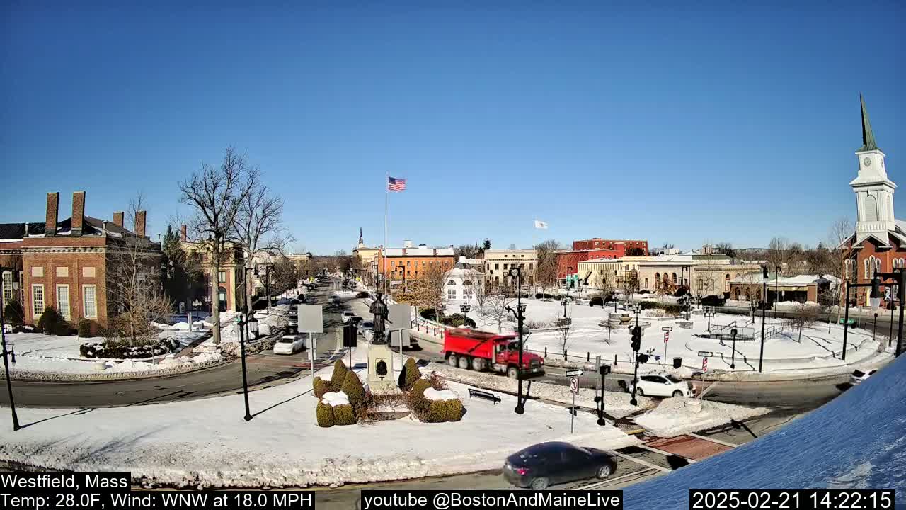 A snow-covered town square with brick buildings, a few cars, and a red dump truck under a sunny sky.