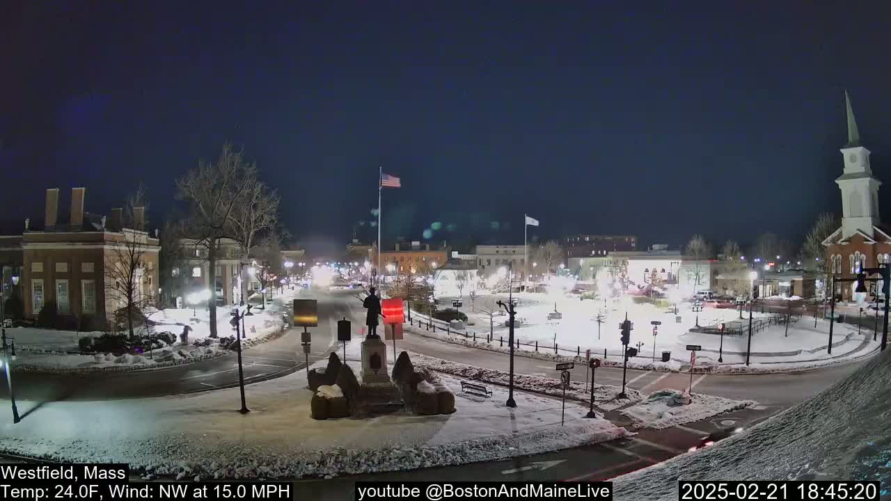 A snow-covered town square at night, illuminated by streetlights, features a statue at its center with buildings and a church visible in the background under a dark sky with a northwest wind.