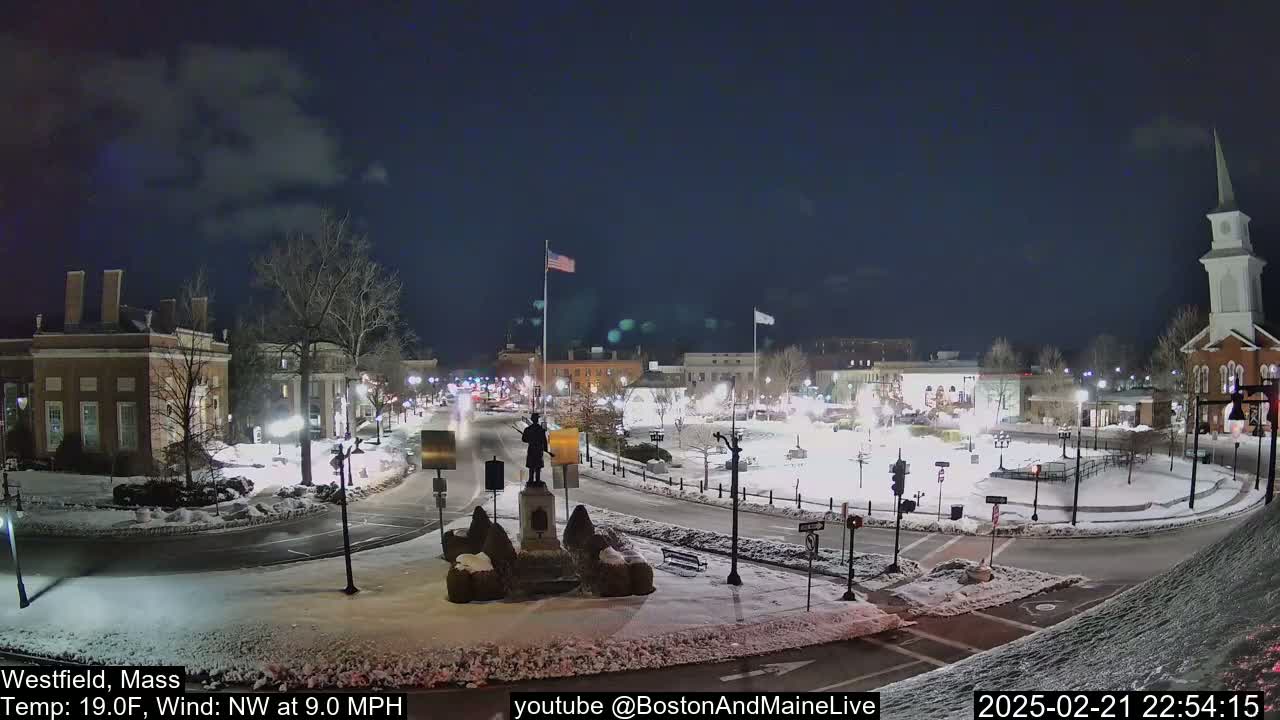 A snow-covered town square at night, illuminated by streetlights, features a monument in the center, surrounded by buildings and a church steeple in the background under a partly cloudy sky.