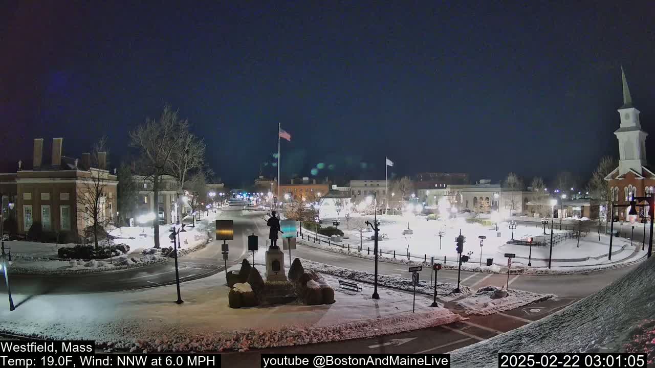 A snow-covered town square at night, illuminated by streetlights, features a statue in the center and several buildings surrounding it, under a clear, dark sky.