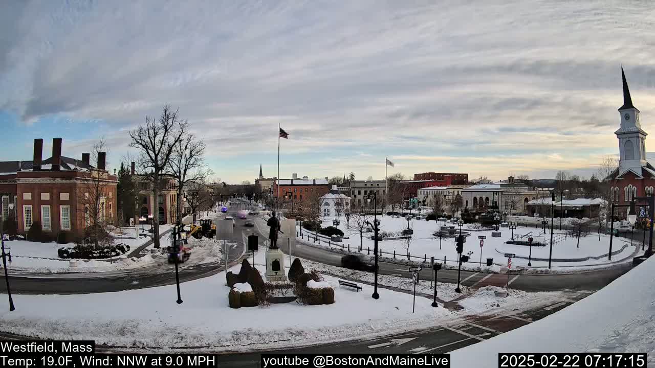 A snow-covered town square with brick buildings, a statue, and a church steeple under a partly cloudy sky.
