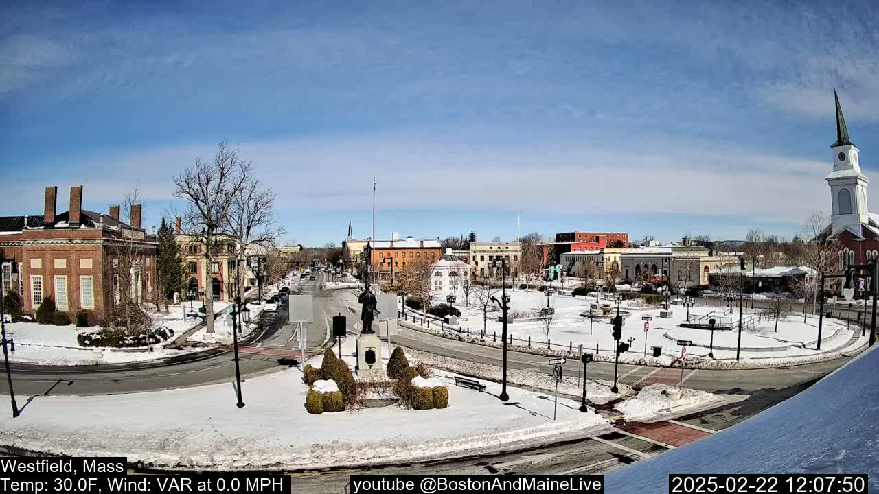 A snow-covered town square with brick buildings, a statue, and a church steeple under a sunny sky.