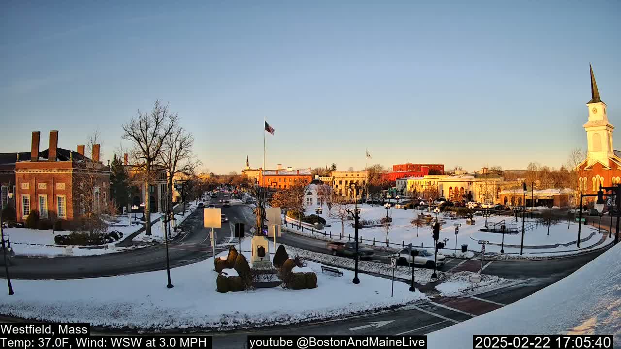 A snow-covered town square with brick buildings, a church steeple, and a monument in the center, under a clear, twilight sky.